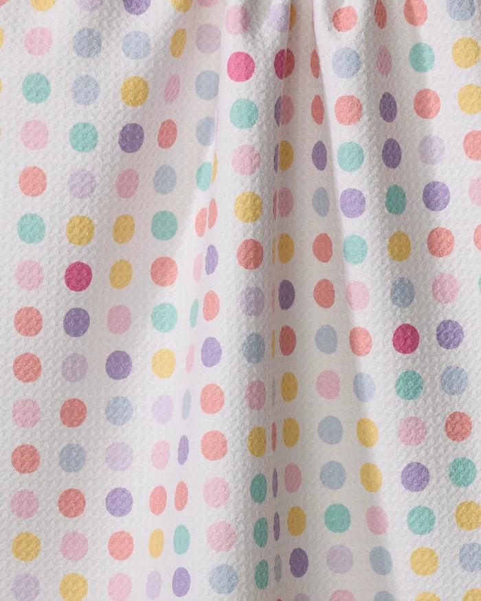 The "Lots of Dots" tea towel by Rebecca Bobko features a white textured fabric with pastel polka dots in pink, yellow, orange, green, blue, and purple. Slight gathering adds gentle folds and subtle shadows.