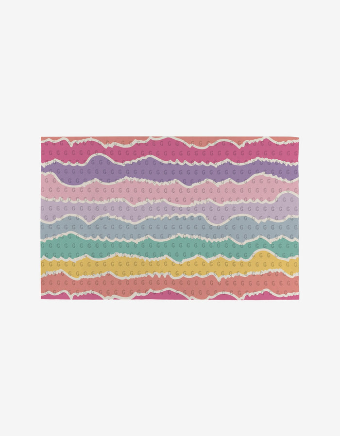 The Rebecca Bobko Lots of Dots mat is a rectangular, reusable mat with wavy pastel stripes in pink, purple, blue, green, and yellow—each stripe features repeating “G”s and a torn-paper texture. An eco-friendly alternative to paper towels.
