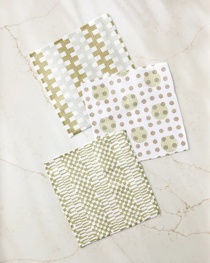 Three absorbent Lotty dishcloths by Studio Jeannot, in green and beige, lie on marble. The set features stylish geometric designs: checks, dots with circles, and zigzags.