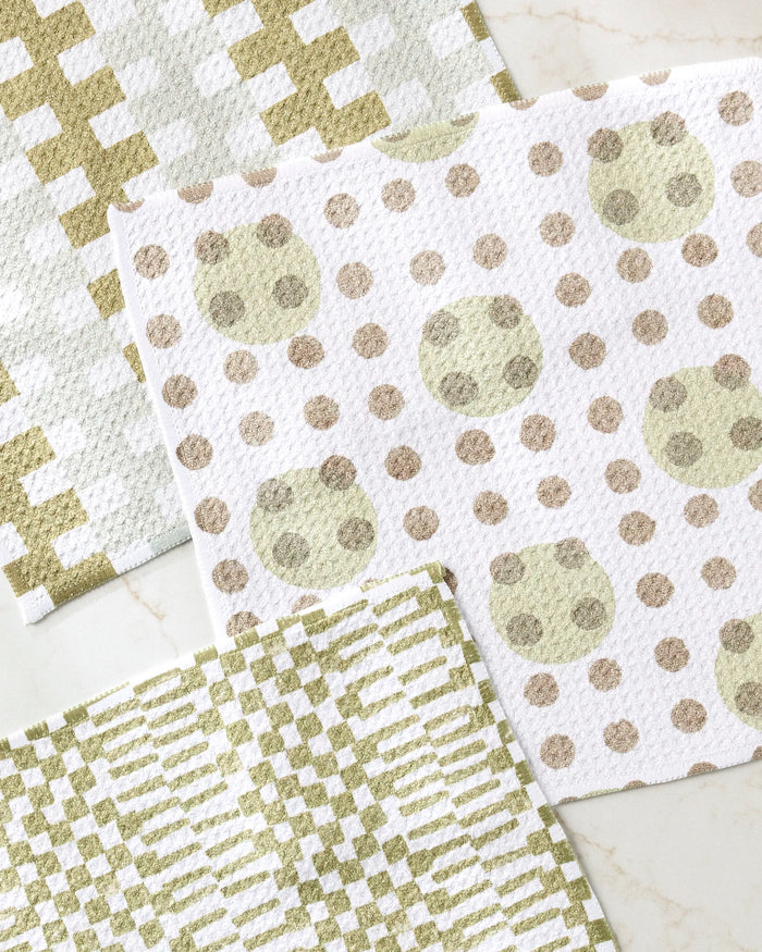 The Studio Jeannot Lotty set features three recycled dishcloths with geometric patterns—gold/white crosses, gold/beige dots, and green/white stripes—stylishly arranged on a light surface.