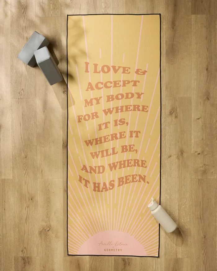 The Love and Acceptance Affirmation yoga mat by Arielle Estoria features yellow with pink rays and the quote “I love & accept my body for where it is, where it will be, and where it has been.” Nearby: grip towel, gray block, white water bottle.