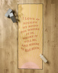 The Love and Acceptance Affirmation yoga mat by Arielle Estoria features yellow with pink rays and the quote “I love & accept my body for where it is, where it will be, and where it has been.” Nearby: grip towel, gray block, white water bottle.