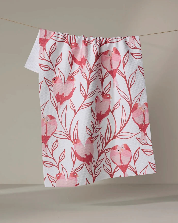 The Love Birds kitchen towel by Hufton Studio, featuring a pink bird-on-branches pattern on white recycled fabric, hangs draped over a clothesline against a plain beige background.