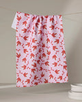 The Love Blooms tea towel by Charlie Rowan Designs, with its red and white floral pattern on pink, hangs on a clothesline brightening the space, while stacked white dishes rest nearby.