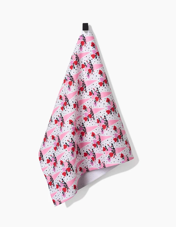 The Love Bouquet tea towel by Charlie Rowan Designs, featuring pink, red, black, and white abstract shapes and dots, hangs from a black hook against a plain white background.