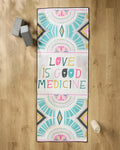 The Love is Good Medicine yoga mat by Jessica Swift features vibrant geometric patterns and the phrase in multicolored letters, accompanied by a non-slip towel, gray yoga blocks, and a water bottle on a wooden floor.
