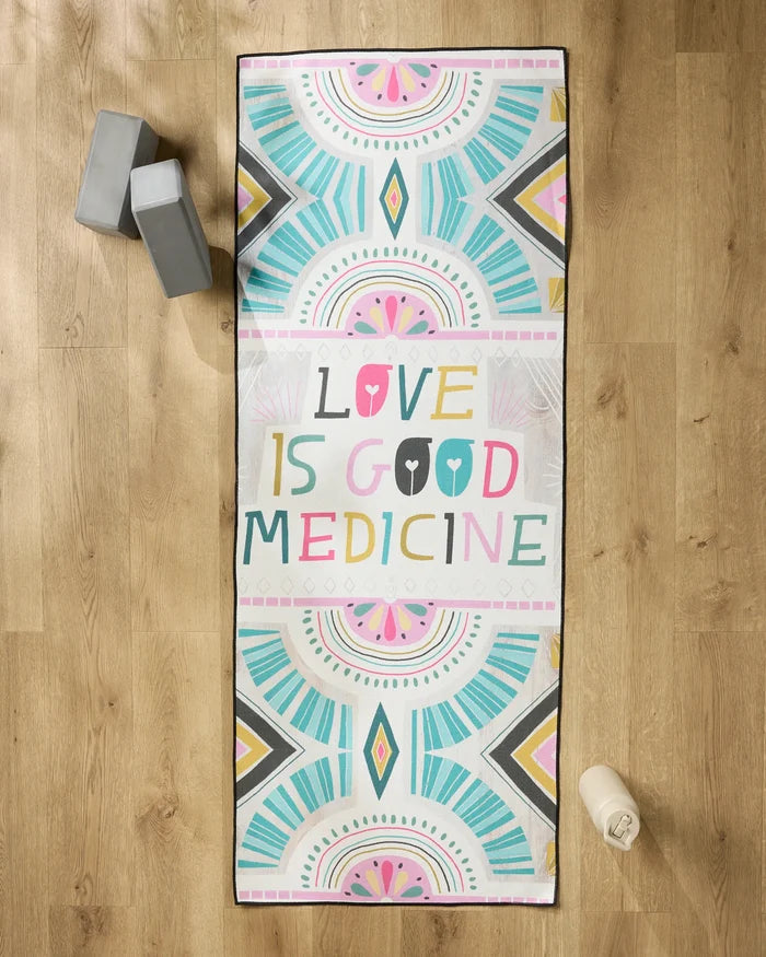The Love is Good Medicine yoga mat by Jessica Swift features vibrant geometric patterns and the phrase in multicolored letters, accompanied by a non-slip towel, gray yoga blocks, and a water bottle on a wooden floor.