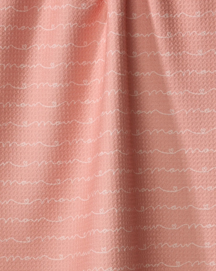 A close-up of the Love of Mom kitchen towel by Sarah Reed, featuring a soft pink, recycled fabric with white wavy lines and repeating cursive text on its gently textured, slightly gathered surface.