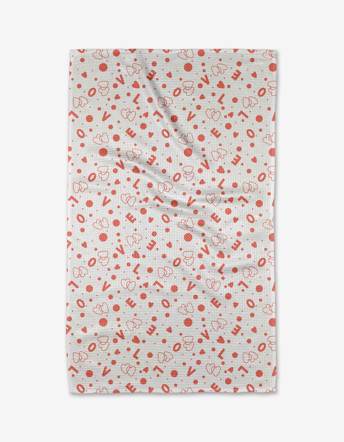 The Love Party kitchen towel by Charlie Rowan Designs is white, made from recycled materials, and features red hearts, dots, and abstract shapes. It appears slightly wrinkled and is shown against a plain white background.