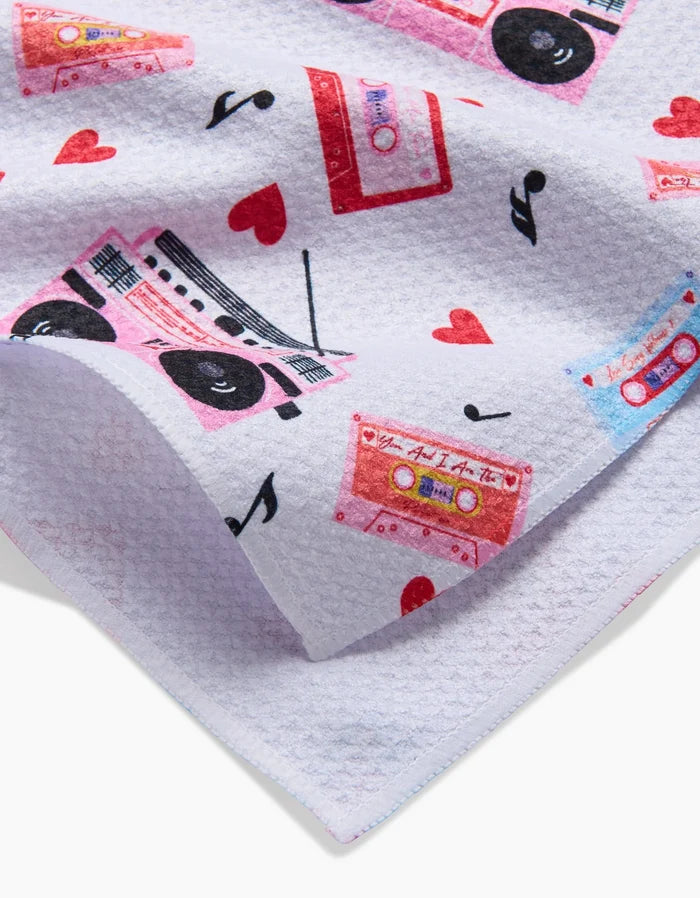A close-up of the Love Playlist tea towel by Vaishali Design Studio, showcasing a textured white fabric with colorful cassette tapes, boomboxes, red hearts, and musical notes.