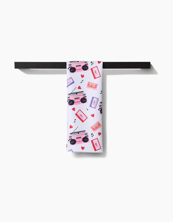 The Love Playlist recycled kitchen towel by Vaishali Design Studio, with cassette tapes, boomboxes, music notes, and red hearts prints, hangs neatly over a black towel bar against a white background.