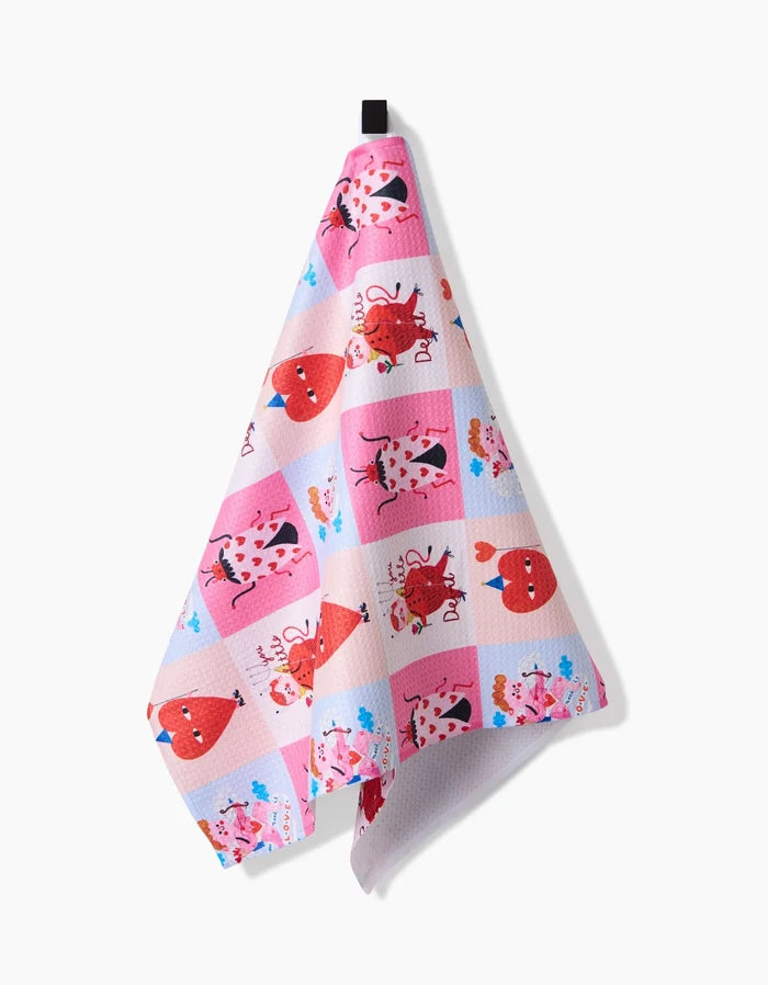 The Love Squares tea towel by Daria Solak, made from recycled materials and featuring pink and white checks with cartoon animal faces and hearts, hangs on a black hook against a plain white background.