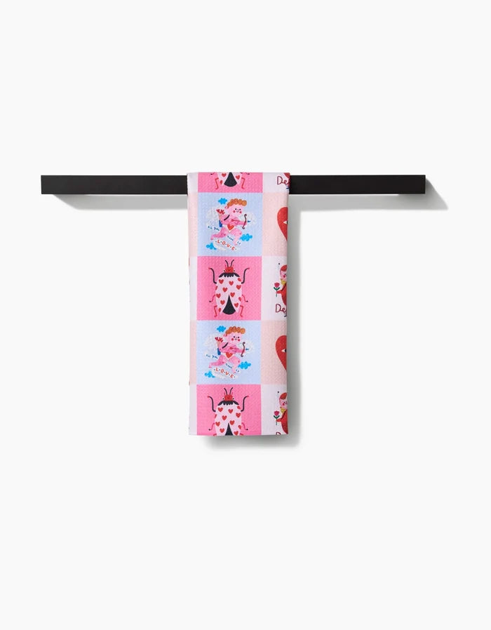 The Daria Solak Love Squares tea towel, featuring colorful cow and heart motifs and crafted from recycled materials, is neatly folded over a black towel bar against a white background.