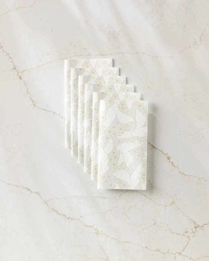 Six folded, reusable GEOMETRY "Love Takes Flight" paper towels, featuring a subtle light beige bird and branch pattern, are neatly arranged in a row on white marble with light brown veining.
