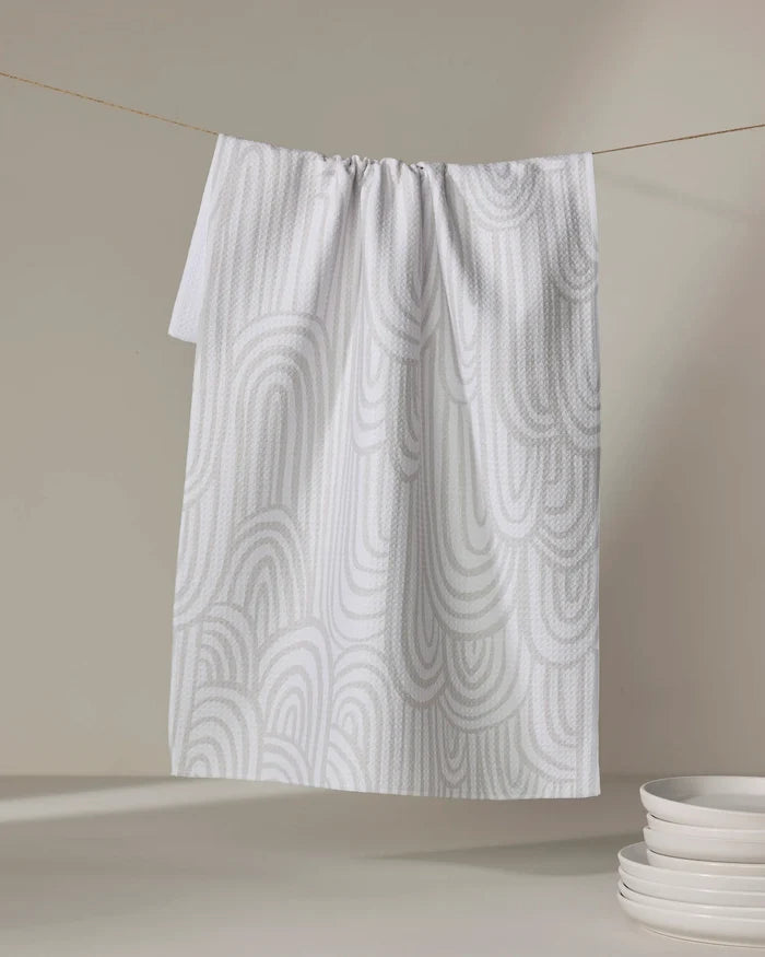 A recycled kitchen towel, Lovers Arc by Hufton Studio, in white with a light gray abstract wave pattern hangs on a line. A small stack of white bowls sits on a neutral surface in the lower right corner.
