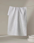 A recycled kitchen towel, Lovers Arc by Hufton Studio, in white with a light gray abstract wave pattern hangs on a line. A small stack of white bowls sits on a neutral surface in the lower right corner.
