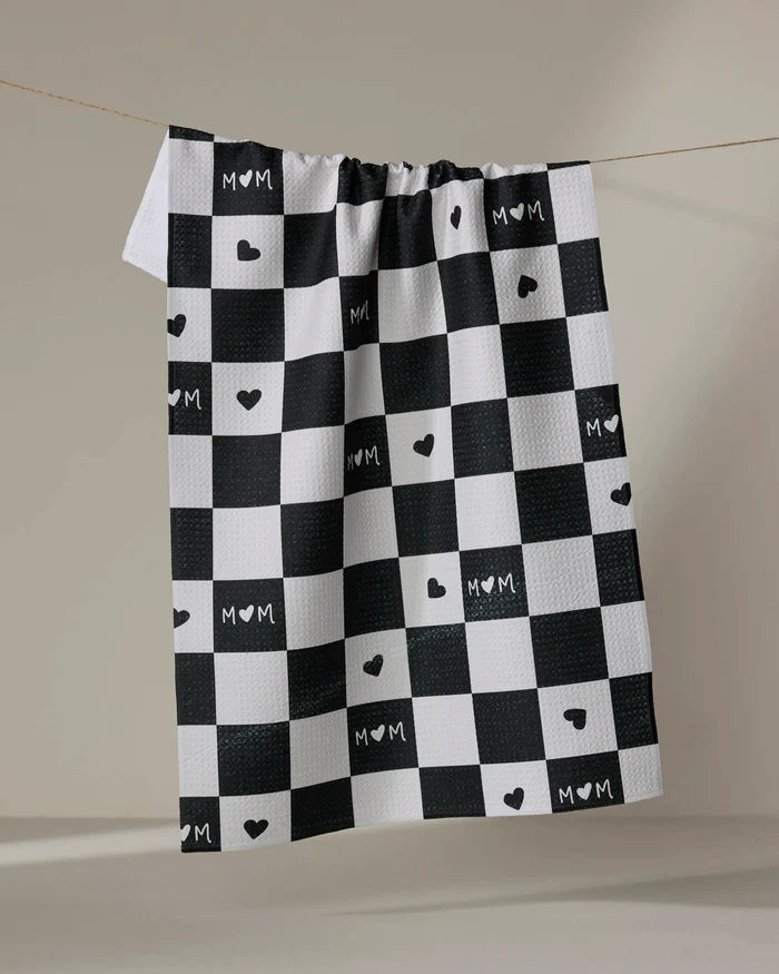 The GEOMETRY "Loving" tea towel features a black and white checkered pattern with MOM and heart symbols on some squares, hanging on a line against a neutral background.