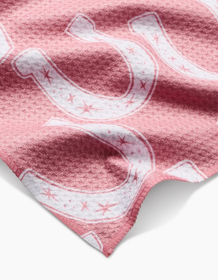 A close-up of the Aubrey Fairchild Lucky Horseshoe bar towel shows its textured pink Geoweave fabric with large white horseshoes and small stars. Made from recycled material, the soft towel has visible folds and shadows.
