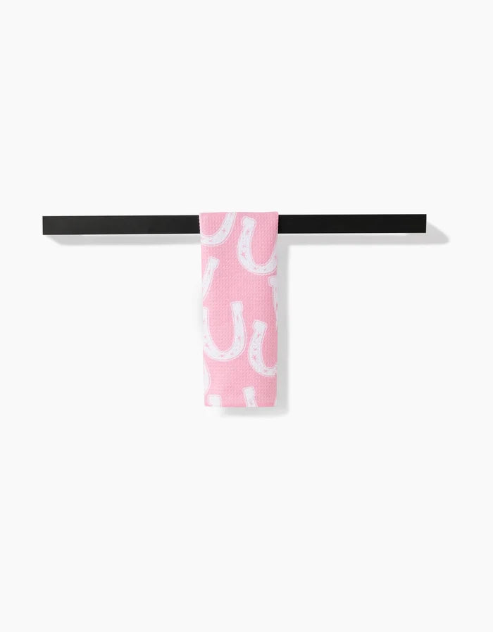 The Lucky Horseshoe bar towel by Aubrey Fairchild, featuring a white horseshoe pattern on pink Geoweave fabric, hangs over a black bar against a plain white background.