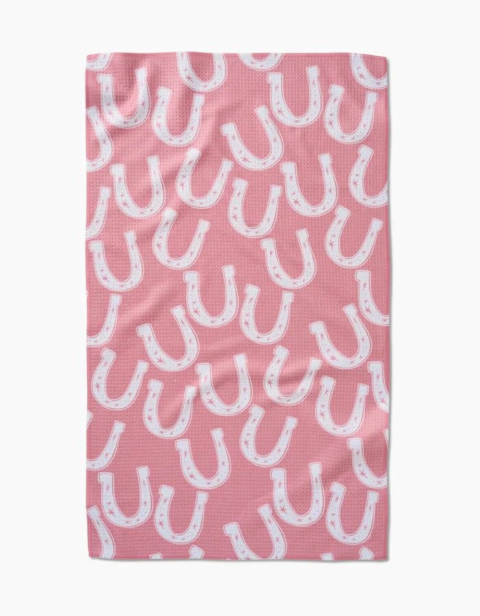 The Lucky Horseshoe tea towel by Aubrey Fairchild features a pink Geoweave fabric with a repeating pattern of white horseshoes outlined in black and accented with small pink stars.