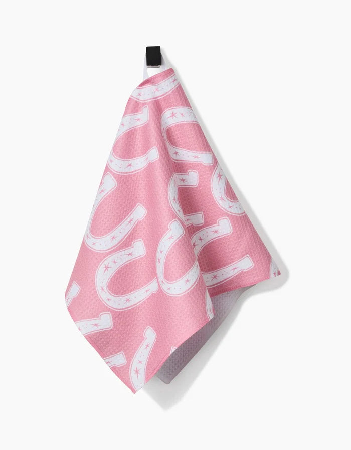 The Lucky Horseshoe Bar Towel by Aubrey Fairchild, made from Geoweave fabric, features a pink design with white horseshoes and small stars. It hangs from a black loop against a white background.