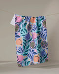 The Lucky Pomegranate kitchen towel by Masha Volnova, made from recycled materials, features a vibrant abstract fruit and leaf design in pink, blue, green, and yellow, hanging against a plain beige background.