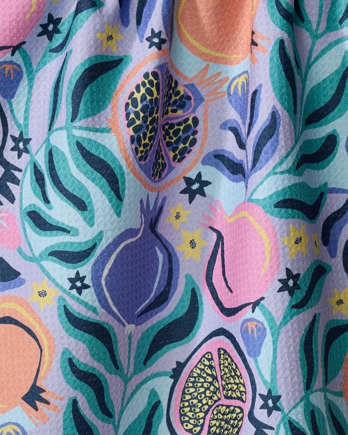 The Lucky Pomegranate kitchen towel by Masha Volnova features a lively, stylized pomegranate and leaves pattern in vibrant purple, pink, teal, green, and yellow on textured recycled fabric.