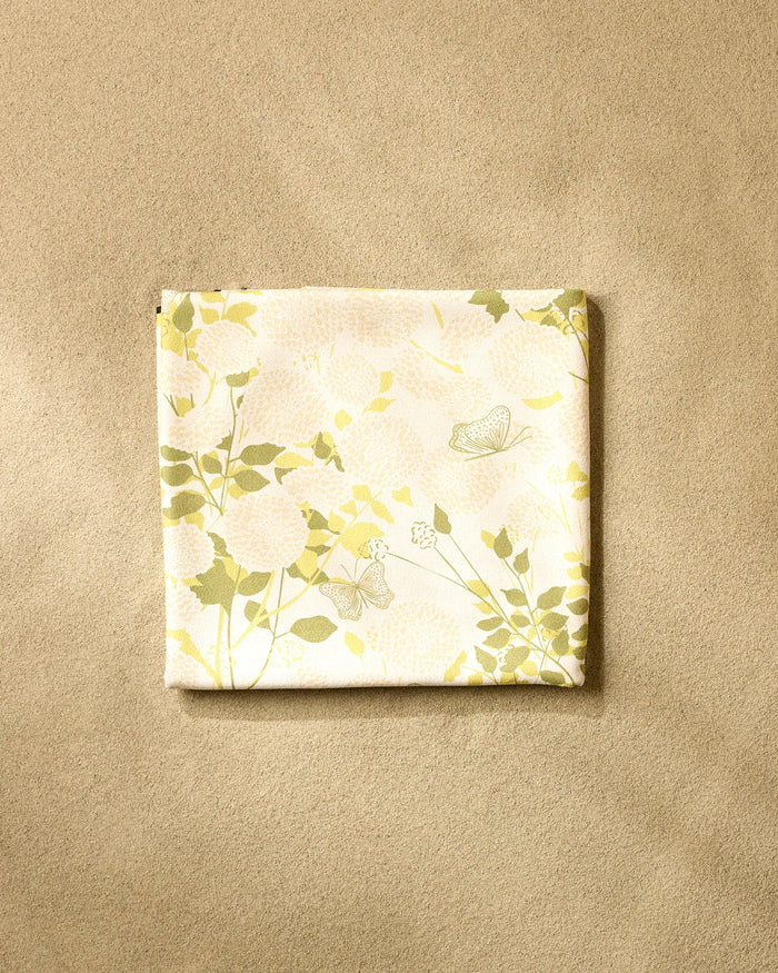 The Lush Garden beach blanket by Second Nature Studio, made from recycled materials and adorned with a soft yellow and green floral-butterfly design, is shown folded on a beige textured surface.