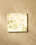 The Lush Garden beach blanket by Second Nature Studio, made from recycled materials and adorned with a soft yellow and green floral-butterfly design, is shown folded on a beige textured surface.