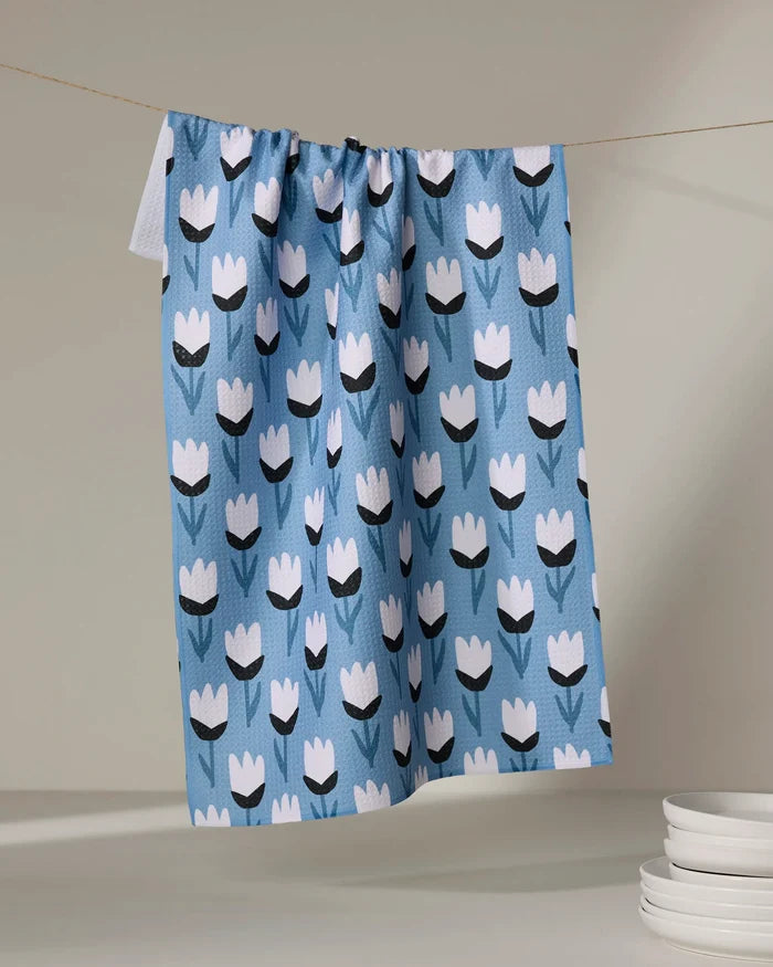 The Lush Meadow tea towel by Anette Heiberg, made from recycled materials and featuring a blue design with white and black tulips, hangs on a clothesline with white plates stacked at the bottom right against a neutral background.