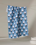 The Lush Meadow tea towel by Anette Heiberg, made from recycled materials and featuring a blue design with white and black tulips, hangs on a clothesline with white plates stacked at the bottom right against a neutral background.