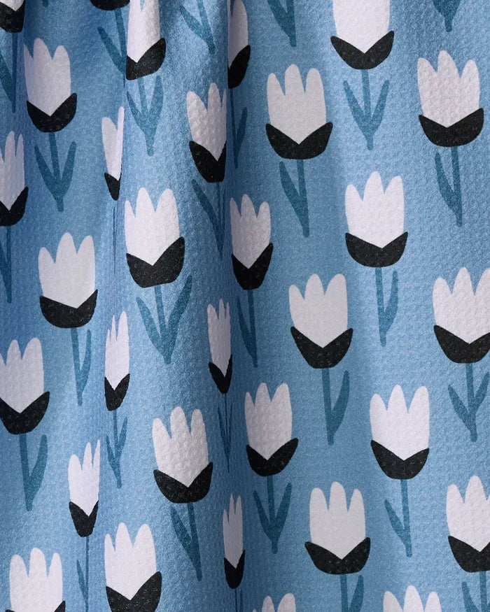 A close-up of the Lush Meadow kitchen towel by Anette Heiberg shows textured light blue fabric with a charming pattern of white tulip flowers, black centers, and blue stems—an elegant touch for any kitchen.