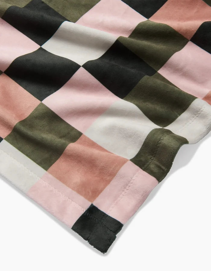 A close-up of the Geometry Luxora blanket, showcasing a large checkered pattern in pink, olive green, black, and white. The soft, slightly wrinkled fabric features edge stitching and is crafted from eco-friendly recycled materials.