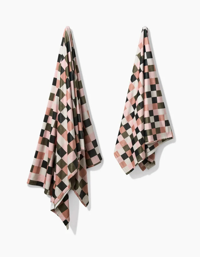 The Geometry Luxora set includes two recycled towels with a geometric checkered pattern in pink, black, white, and olive green, displayed side by side on a white background.