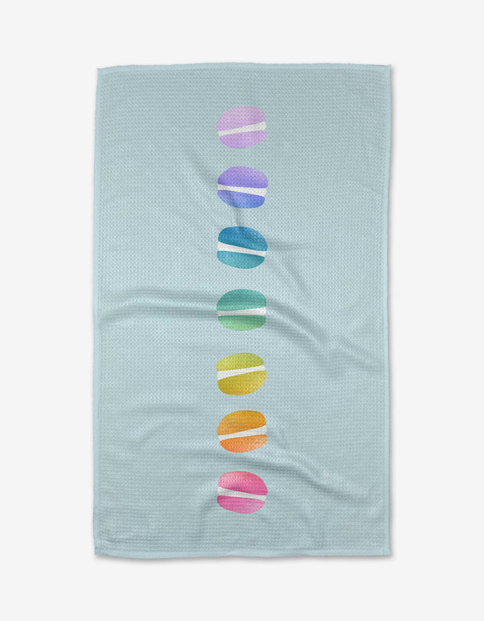The Mac Love Kitchen Tea Towel by Noras Macarons features light blue Geoweave fabric with a centered vertical row of seven colorful, rainbow-hued macaron ovals.