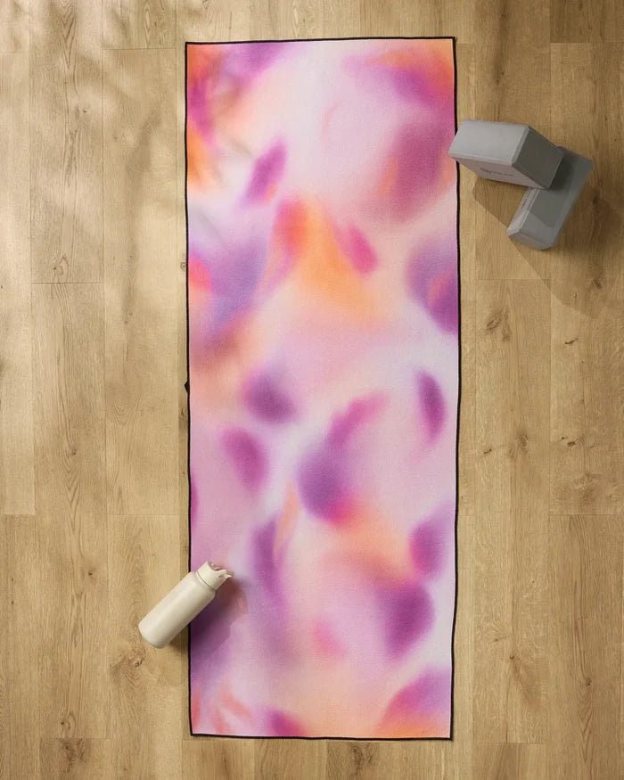 The GEOMETRY Magenta yoga mat, featuring pink, orange, and purple swirls, rests on a wooden floor. A white water bottle sits at the bottom left corner with two gray yoga blocks and a non-slip towel at the top right.