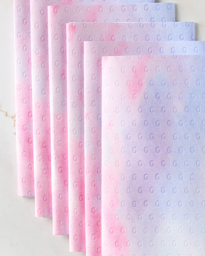 Six pastel ombré Magic In The Sky napkins by Stefanie Bales, featuring a small G-letter pattern and hues that fade from pink to purple and blue, are arranged overlapping. These reusable towels offer an eco-friendly kitchen alternative.