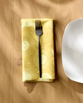 The Jessica Swift "Magical Moments" yellow reusable cloth napkin with sun and star patterns holds a black fork and sits beside a white square plate on a wooden table.