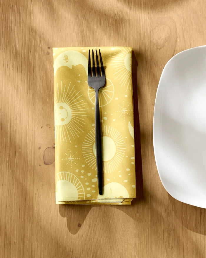 The Jessica Swift "Magical Moments" yellow reusable cloth napkin with sun and star patterns holds a black fork and sits beside a white square plate on a wooden table.