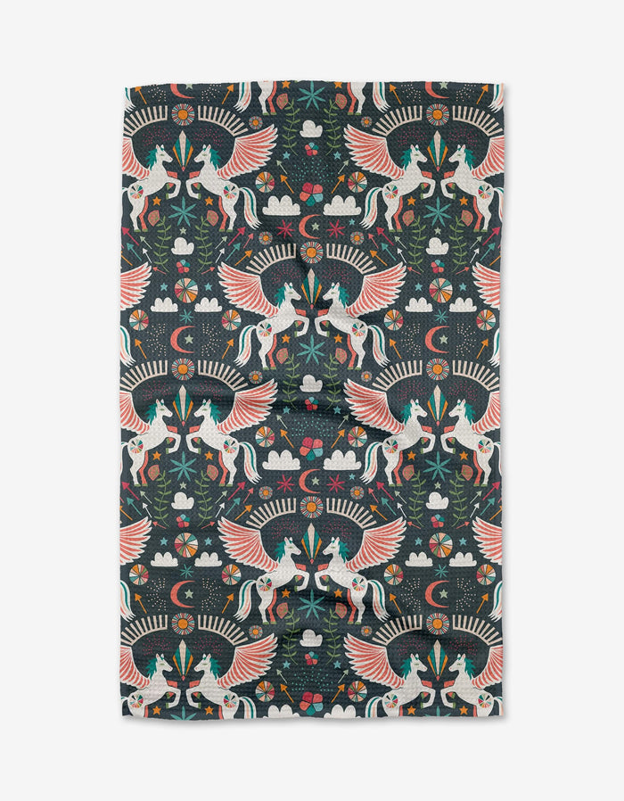 Jessica Swift’s Magical Moments kitchen towel showcases colorful winged unicorns, flowers, stars, clouds, and foliage in a symmetrical folk-art style on a dark background.