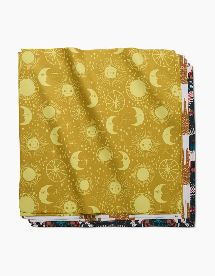 A stack of folded fabric squares, with Jessica Swift’s Magical Moments on top—a yellow print with smiling suns, moons, and stars—makes perfect eco-friendly napkins for your Dinner Napkin Set. Other colorful patterns peek out below.