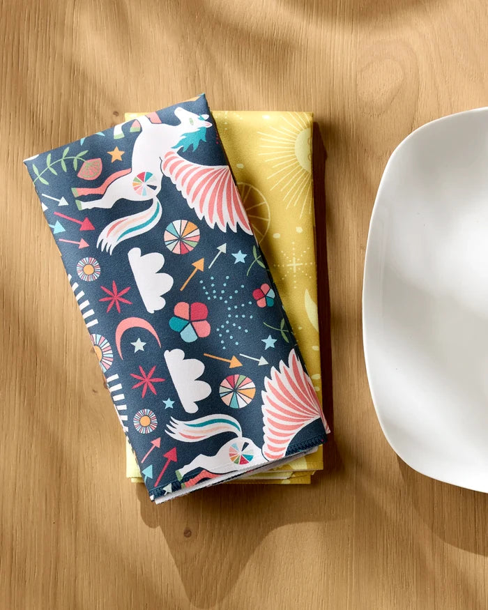 Two folded "Magical Moments" eco-friendly napkins by Jessica Swift rest on a wooden table; the top shows colorful birds and geometric patterns, while the yellow bottom napkin displays sun motifs.