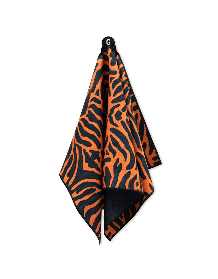 A GEOMETRY Magna - Tiger fitness towel, featuring an orange and black tiger stripe pattern and made from recycled materials, hangs from a hook against a white background.