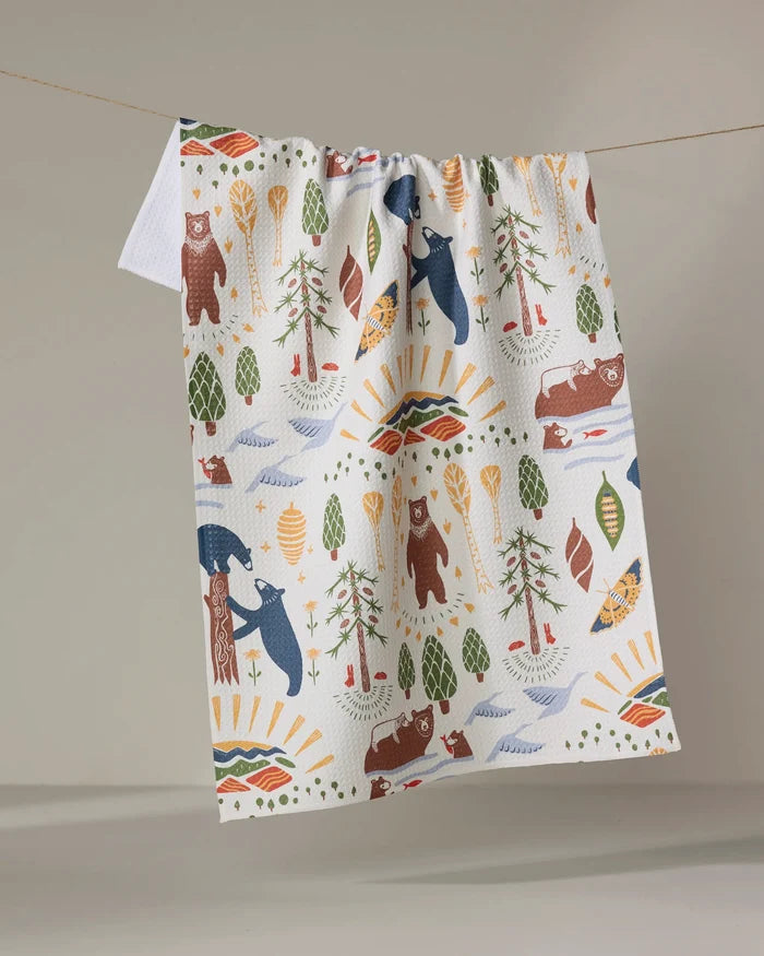 The Mama Bear recycled kitchen towel by Elisabeth Wing, decorated with vibrant wilderness motifs—bears, trees, canoes, fish, paddles, rivers, and suns—hangs on a clothesline against a plain backdrop.