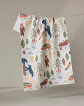 The Mama Bear recycled kitchen towel by Elisabeth Wing, decorated with vibrant wilderness motifs—bears, trees, canoes, fish, paddles, rivers, and suns—hangs on a clothesline against a plain backdrop.