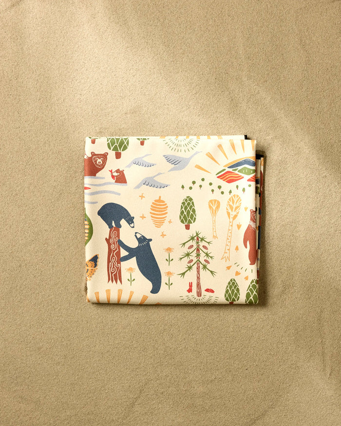 The "Mama Bear" folded beach blanket by Elisabeth Wing, crafted from recycled materials and featuring bears, trees, pinecones, and sun motifs in earthy tones, rests on a sandy beige background.