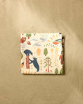 The "Mama Bear" folded beach blanket by Elisabeth Wing, crafted from recycled materials and featuring bears, trees, pinecones, and sun motifs in earthy tones, rests on a sandy beige background.