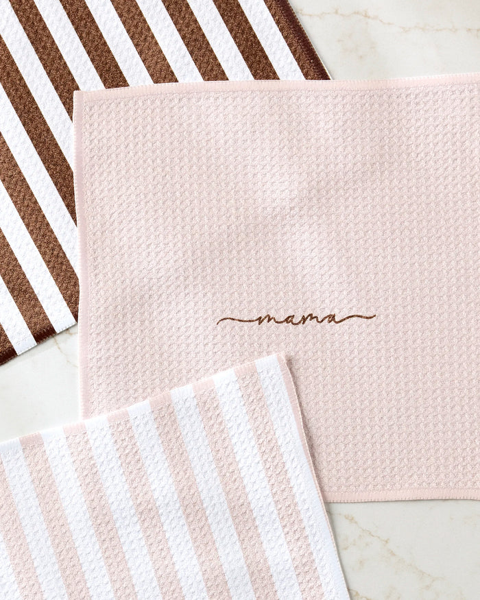 Three Kailee Wright “Mama” dish towels are shown: brown and white stripes, pink and white stripes, and light pink with “mama” embroidered in brown, all displayed on a light marble surface.