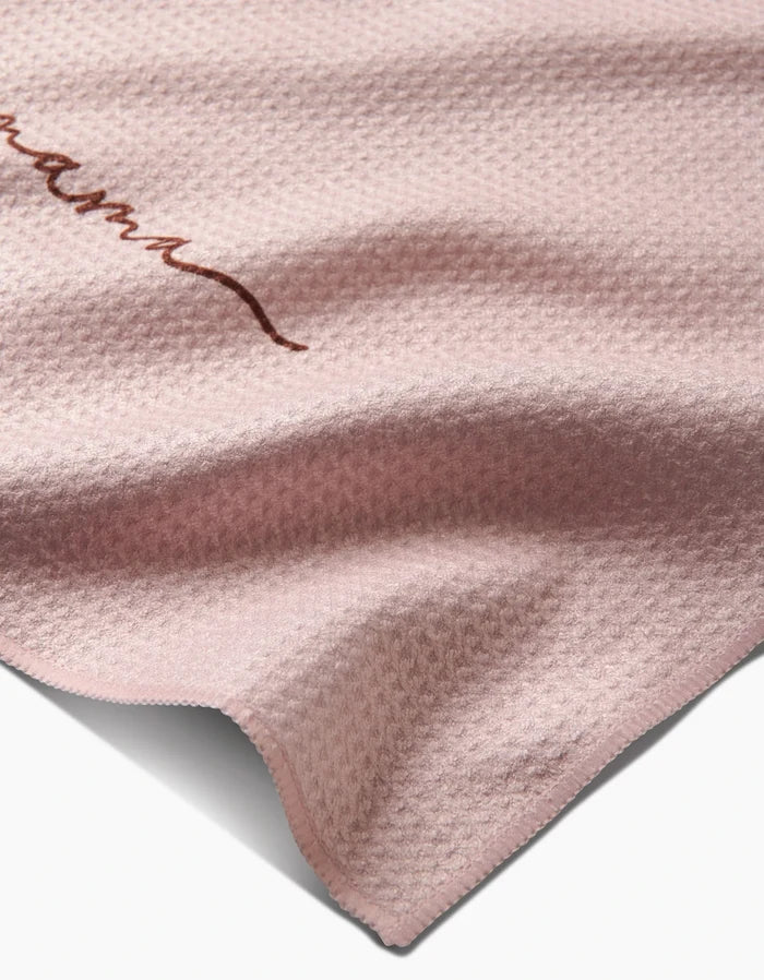 A close-up of the Kailee Wright Mama towel, in textured light pink Geoweave fabric with a subtle wavy pattern and a small Mama embroidery in brown on the upper left side.