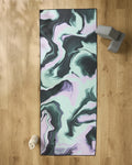 The GEOMETRY Marble yoga mat, featuring a black, mint green, and lavender marbled design, rests on a wooden floor alongside a non-slip towel, water bottle, and gray yoga blocks.